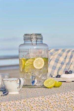 Saftdispenser i klart glas med tappehane fra Ib Laursen stående på bord på stranden