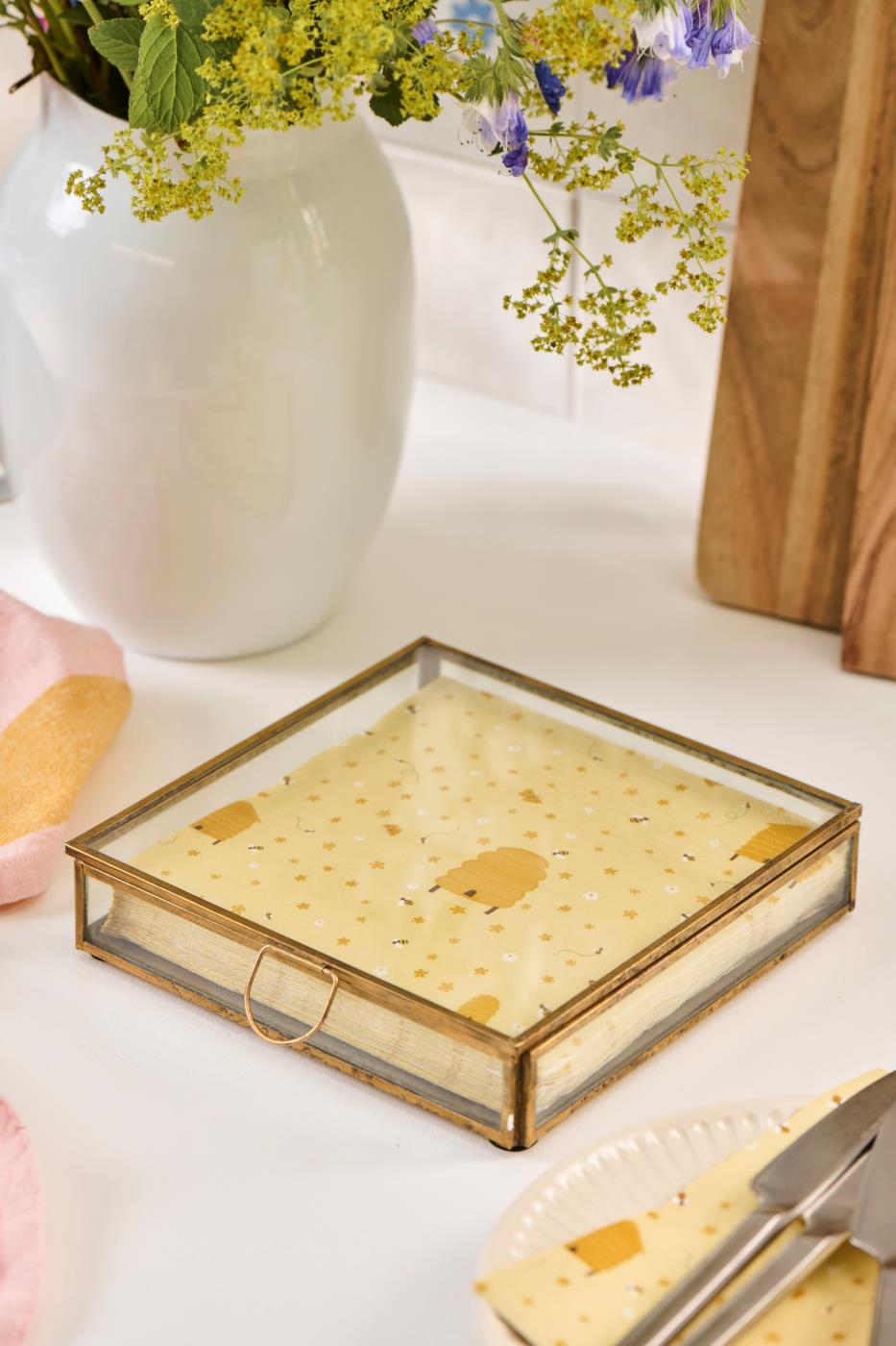 Solitaire glass box with a gold-colored frame and napkins on a light table next to a vase of flowers