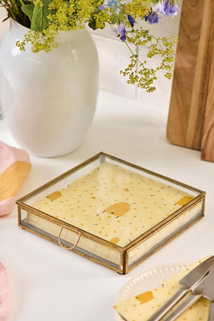 Solitaire glass box with a gold-colored frame and napkins on a light table next to a vase of flowers
