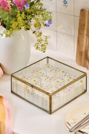 Solitaire glass box with a gold-colored frame and napkins on a light table next to a vase of flowers